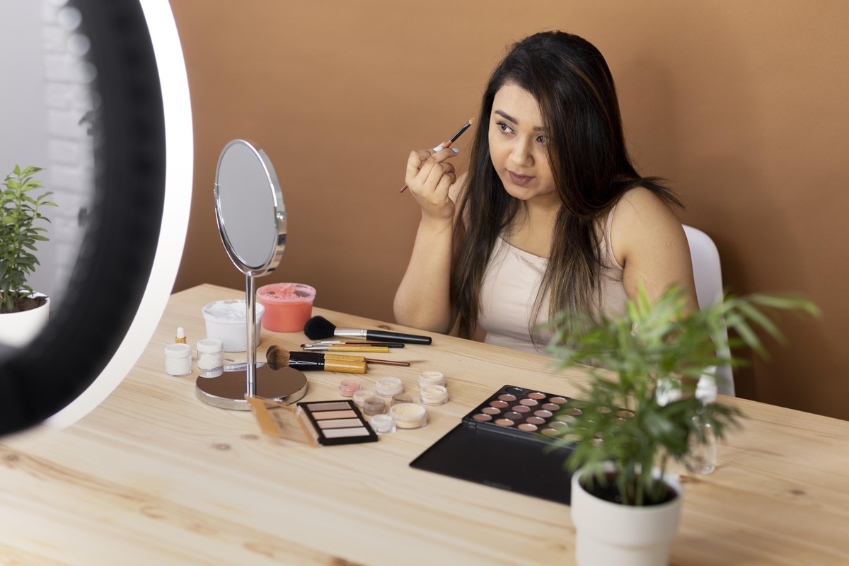 A woman sits at a wooden desk applying makeup while filming a tutorial. Various makeup products and brushes are spread out on the table, and a small mirror is in front of her. A ring light is visible in the foreground, illuminating the scene. There is a potted plant on the desk, adding a natural touch to the setting , foundation.