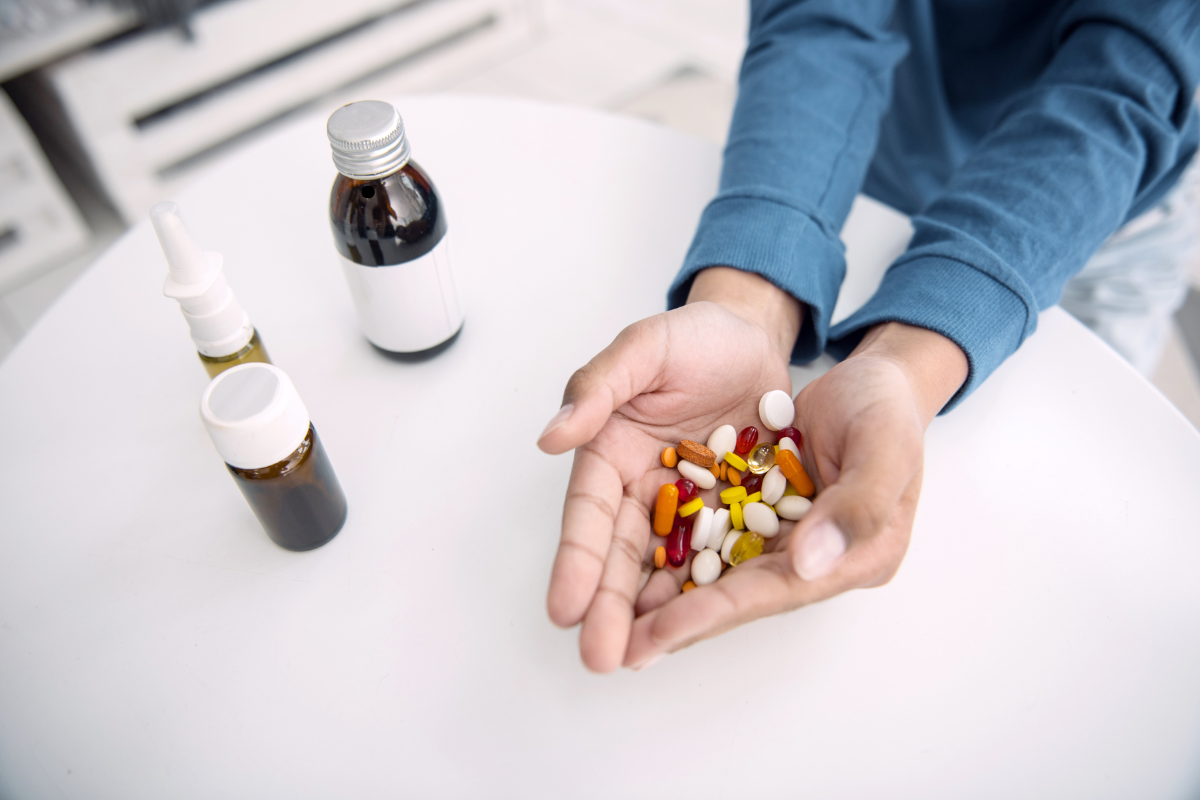 A person holding a variety of pills and capsules in their hands, with several bottles of medication on a white table.